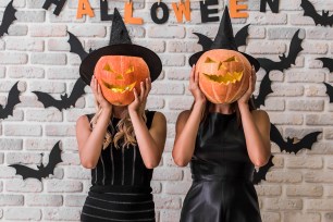 Beautiful girls in black dresses and witch hats are holding scary pumpkins, on background decorated for Halloween